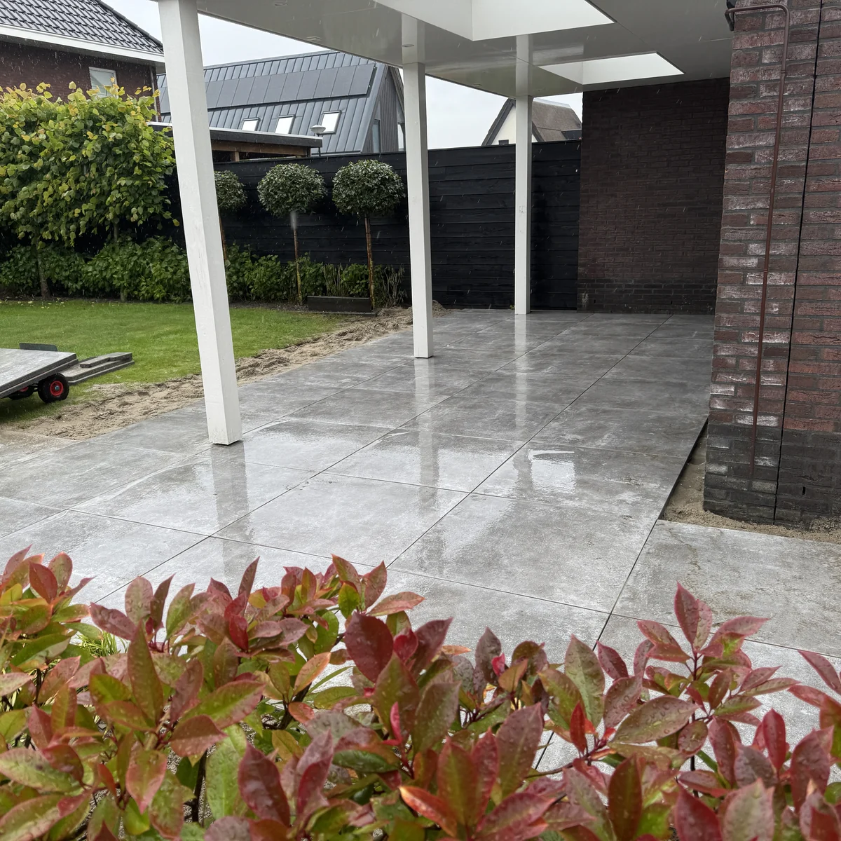 Terras van grote keramische tegels onder een witte overkapping, met photinia-haag op de voorgrond